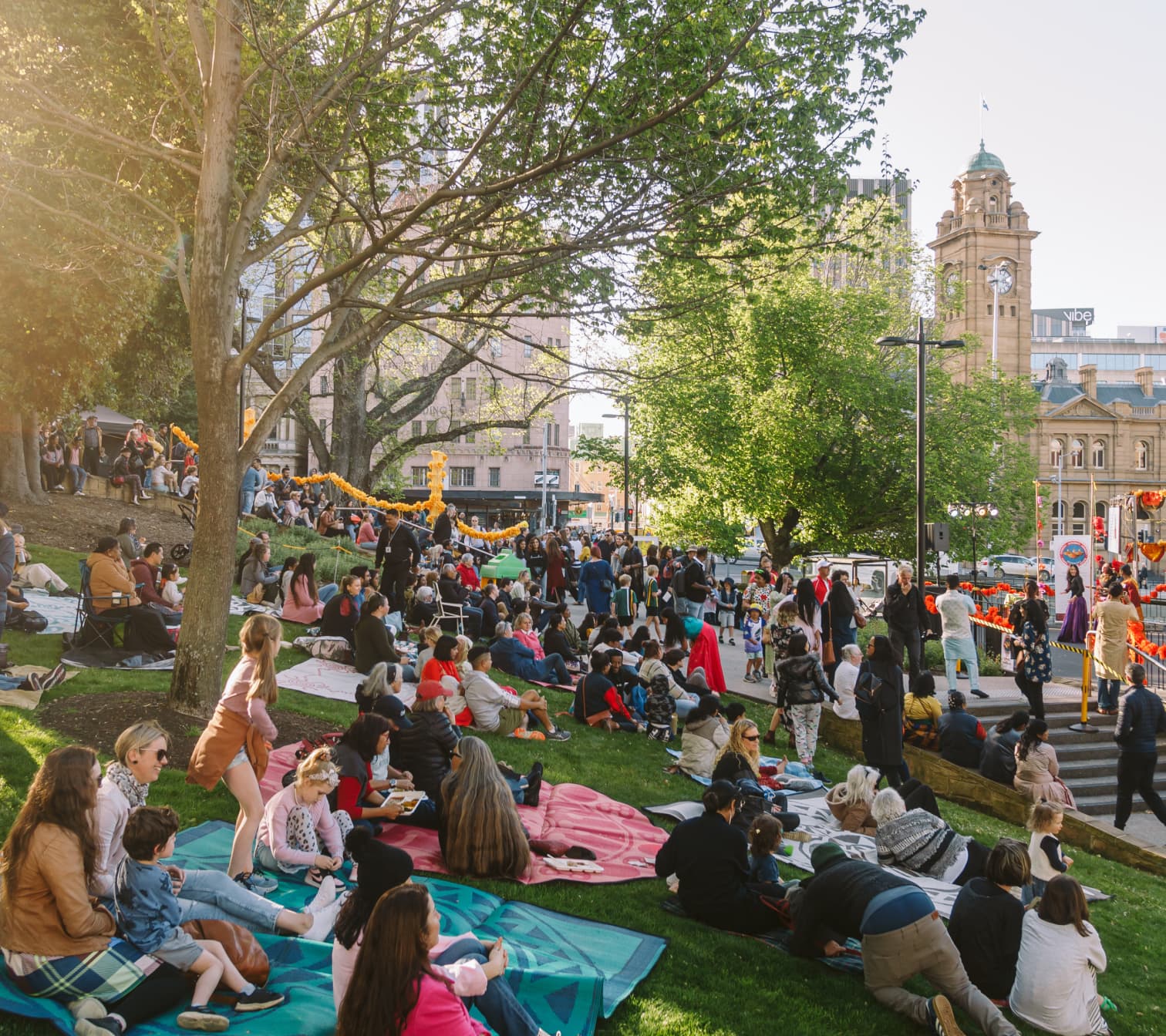 People gather in Franklin Square to enjoy live entertainment. Families sit on colourful picnic blankets beneath shady trees.